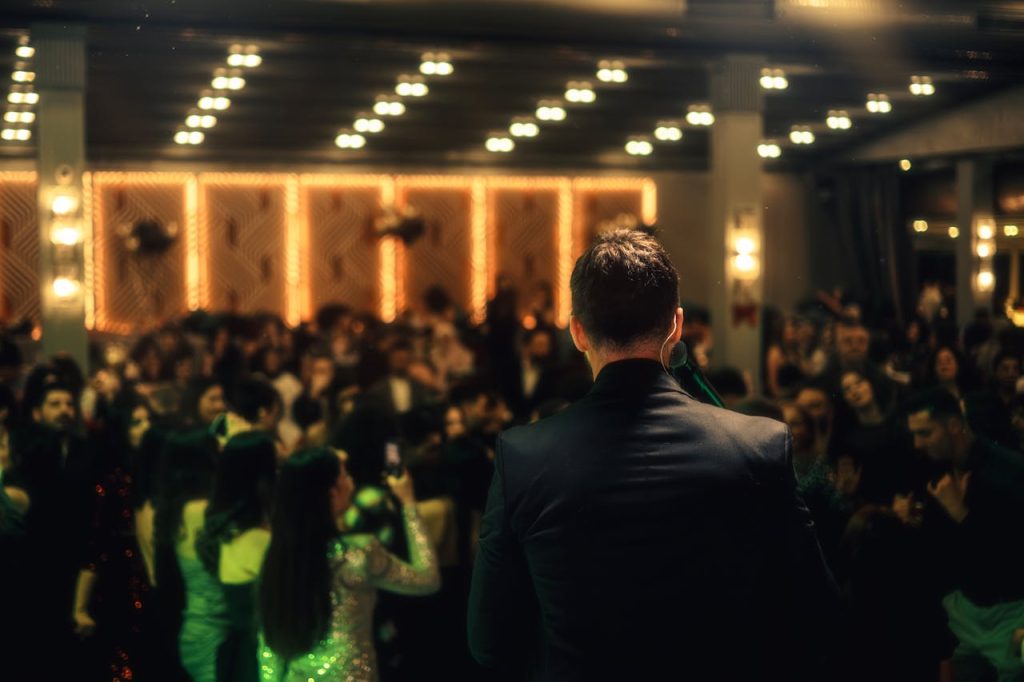 A crowded indoor night event with a man addressing the audience, vibrant lighting enhances the scene.